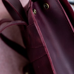 May include: Close-up of a burgundy leather backpack. The bag features a flap closure with brass-colored rivets and adjustable straps. The leather has a slightly distressed appearance, adding to its vintage aesthetic.