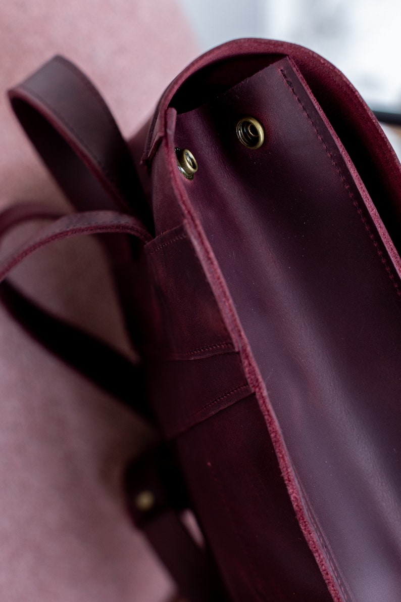 May include: Close-up of a burgundy leather backpack. The bag features a flap closure with brass-colored rivets and adjustable straps. The leather has a slightly distressed appearance, adding to its vintage aesthetic.