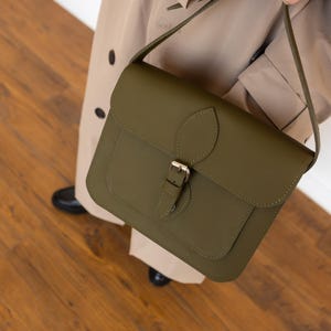 May include: Olive green leather satchel bag with a flap closure and a gold-colored buckle. The bag has a shoulder strap and a small front pocket. The bag is held by a person wearing a tan trench coat.