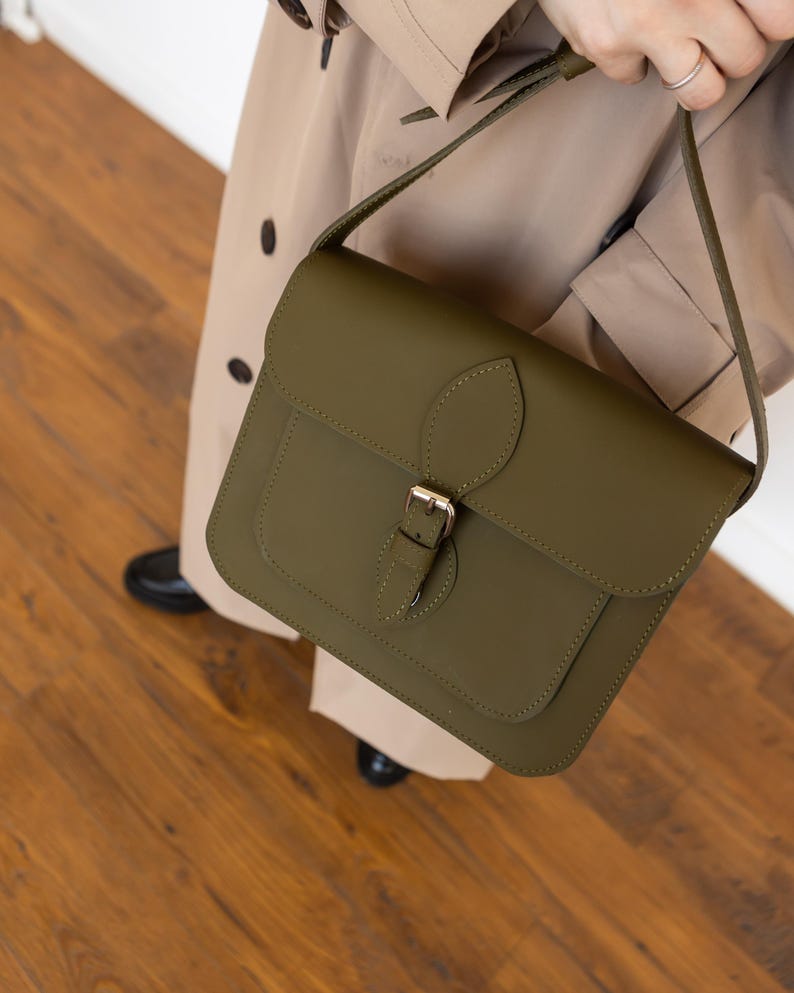 May include: Olive green leather satchel bag with a flap closure and a gold-colored buckle. The bag has a shoulder strap and a small front pocket. The bag is held by a person wearing a tan trench coat.