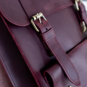 May include: Close-up of a burgundy leather satchel with a flap closure and brass-colored buckles. The bag features a front pocket and adjustable straps. The leather has a rich, textured appearance, and the stitching is visible.