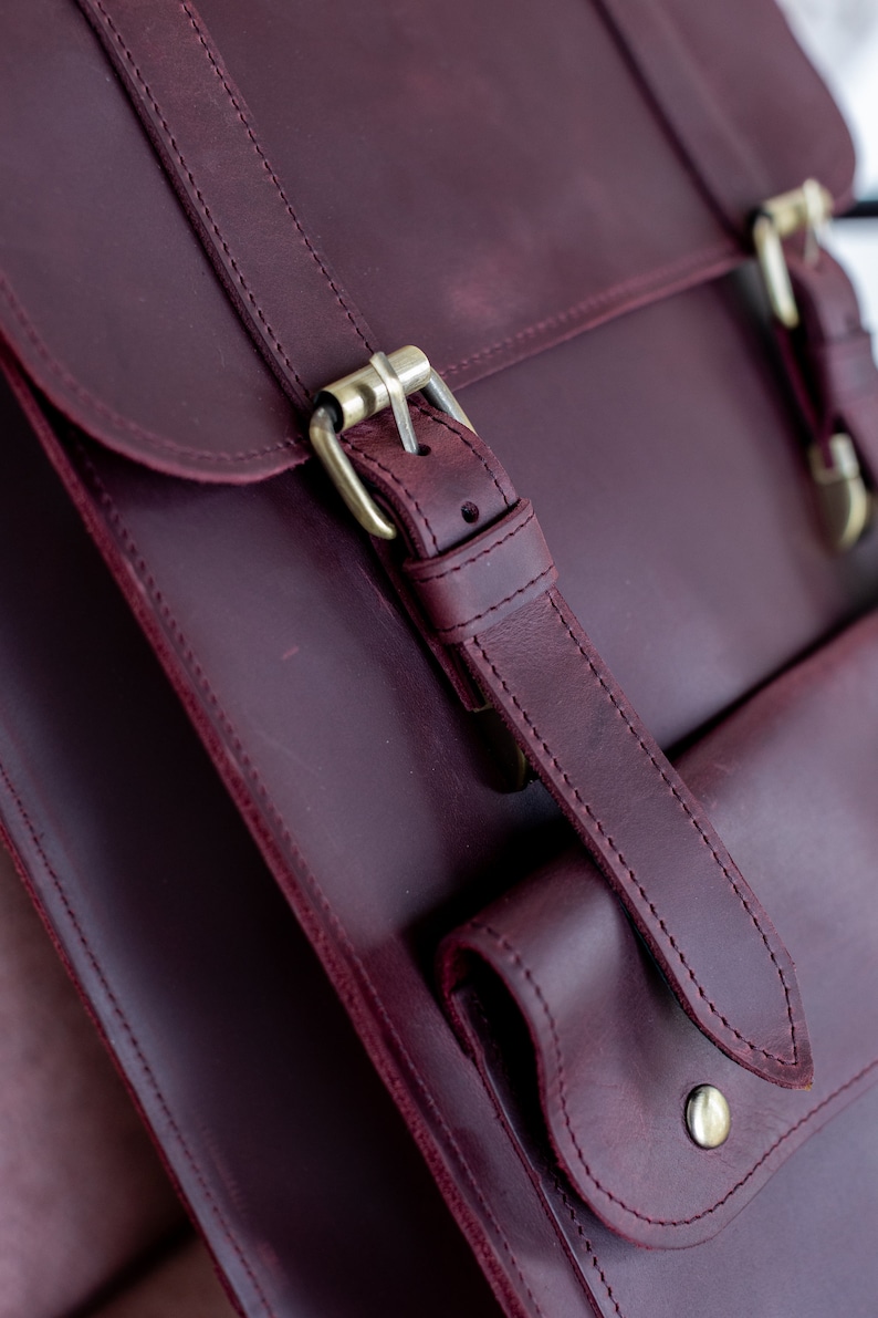 May include: Close-up of a burgundy leather satchel with a flap closure and brass-colored buckles. The bag features a front pocket and adjustable straps. The leather has a rich, textured appearance, and the stitching is visible.