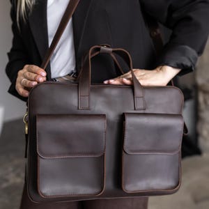 May include: A dark brown leather briefcase featuring two large rectangular front pockets with flap closures, two top handles, and a long adjustable shoulder strap. The bag has a smooth finish and is constructed with visible stitching along the edges.