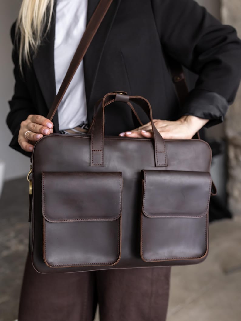 May include: A dark brown leather briefcase featuring two large rectangular front pockets with flap closures, two top handles, and a long adjustable shoulder strap. The bag has a smooth finish and is constructed with visible stitching along the edges.