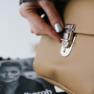 May include: A tan handbag with a silver clasp is being opened. The bag has a rectangular shape and a shiny metal clasp. The background includes a magazine with the text "Peter Lindbergh photography".