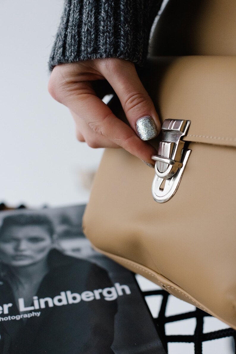 May include: A tan handbag with a silver clasp is being opened. The bag has a rectangular shape and a shiny metal clasp. The background includes a magazine with the text "Peter Lindbergh photography".