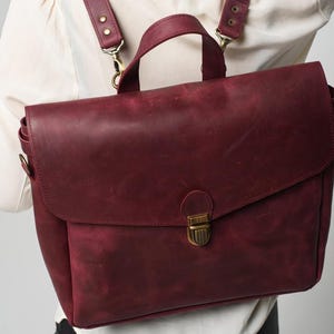 May include: Burgundy leather backpack with adjustable straps and a top handle. The bag has a flap closure with a brass-coloured clasp. The straps are attached with brass-coloured hardware. The bag is worn as a backpack.