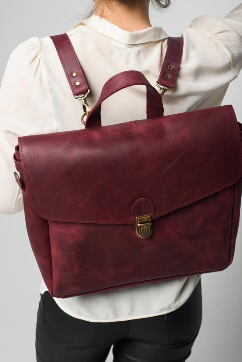 May include: Burgundy leather backpack with adjustable straps and a top handle. The bag has a flap closure with a brass-coloured clasp. The straps are attached with brass-coloured hardware. The bag is worn as a backpack.