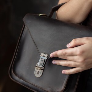 May include: A dark brown leather satchel bag with a rectangular shape and a flap closure. The bag features a metal clasp and a top handle. The leather appears to have a distressed finish, giving it a vintage look. The bag is designed for carrying items.