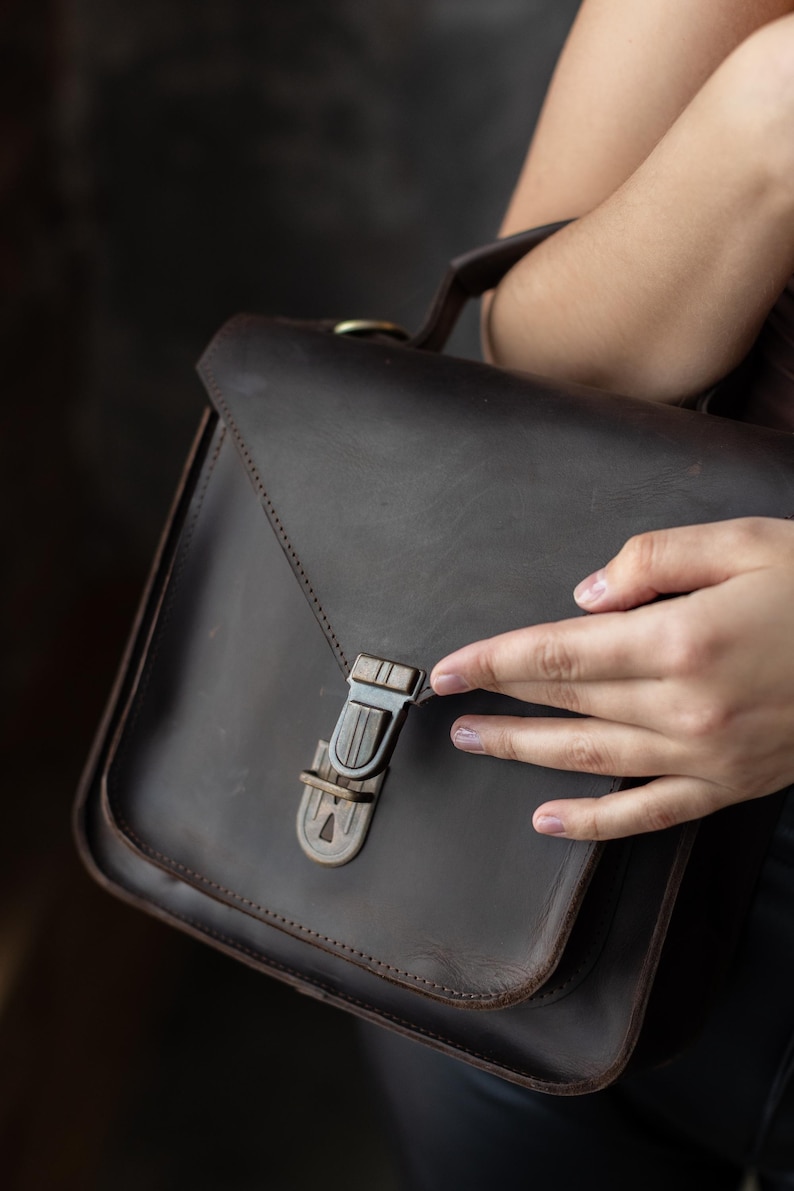 May include: A dark brown leather satchel bag with a rectangular shape and a flap closure. The bag features a metal clasp and a top handle. The leather appears to have a distressed finish, giving it a vintage look. The bag is designed for carrying items.
