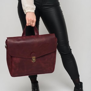 May include: A burgundy leather satchel with a top handle and a flap closure. The bag has a brass-coloured clasp and is held by a person wearing black trousers and boots. The bag is approximately 30 cm wide and 25 cm tall.