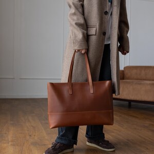 May include: A large, rectangular brown leather tote bag with two long handles. The bag has a simple, minimalist design and is being held by a person wearing a long tan coat and dark blue jeans. The bag appears to be empty.