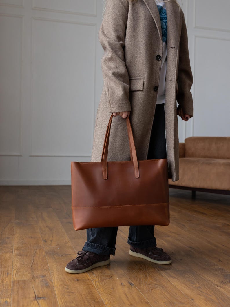 May include: A large, rectangular brown leather tote bag with two long handles. The bag has a simple, minimalist design and is being held by a person wearing a long tan coat and dark blue jeans. The bag appears to be empty.