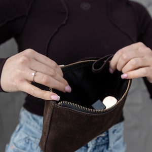 Pu&ograve; includere: Una pochette in camoscio marrone scuro con cerniera dorata &egrave; aperta, rivelando un interno nero. La pochette &egrave; rettangolare e sembra vuota, tranne per un piccolo oggetto rotondo e un piccolo oggetto rettangolare.