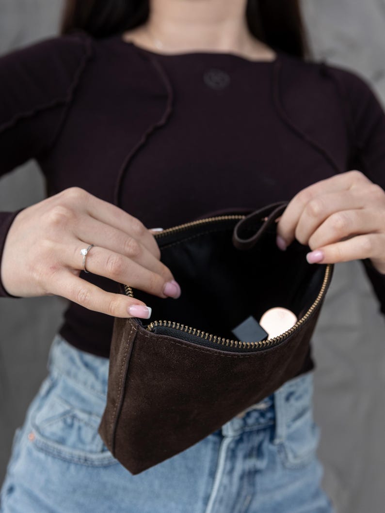 Pu&ograve; includere: Una pochette in camoscio marrone scuro con cerniera dorata &egrave; aperta, rivelando un interno nero. La pochette &egrave; rettangolare e sembra vuota, tranne per un piccolo oggetto rotondo e un piccolo oggetto rettangolare.