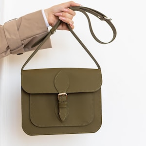 May include: Olive green crossbody bag with a flap closure and adjustable strap. The bag features a front pocket and a gold-colored buckle. The bag is held up by a hand.