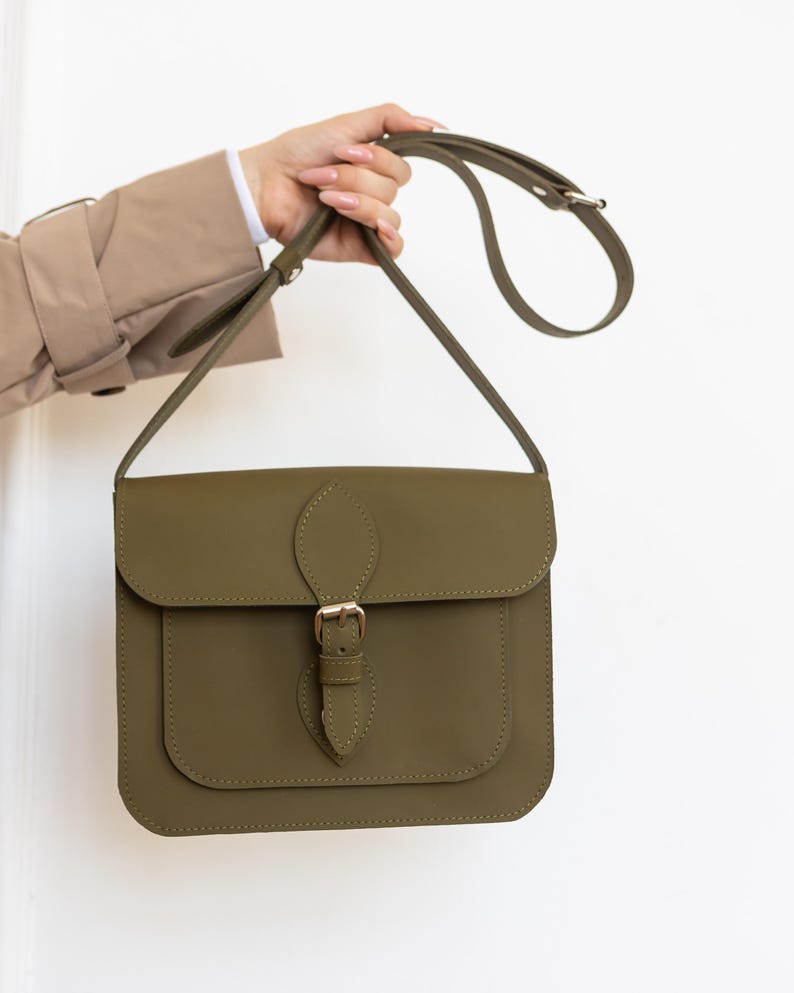 May include: Olive green crossbody bag with a flap closure and adjustable strap. The bag features a front pocket and a gold-colored buckle. The bag is held up by a hand.