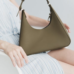 May include: Olive green shoulder bag with a curved design. The bag has a matching strap and silver-tone hardware. The bag is held by a person wearing a blue and white striped dress.