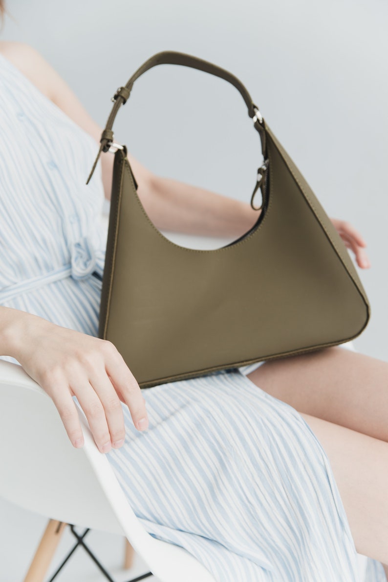 May include: Olive green shoulder bag with a curved design. The bag has a matching strap and silver-tone hardware. The bag is held by a person wearing a blue and white striped dress.