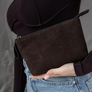 Pu&ograve; includere: Una pochette in camoscio marrone scuro con chiusura a zip e un piccolo manico. La borsa rettangolare &egrave; tenuta su uno sfondo di un top a maniche lunghe viola scuro e jeans.