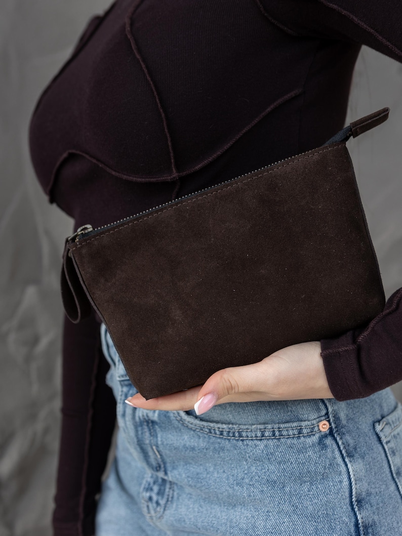 Pu&ograve; includere: Una pochette in camoscio marrone scuro con chiusura a zip e un piccolo manico. La borsa rettangolare &egrave; tenuta su uno sfondo di un top a maniche lunghe viola scuro e jeans.