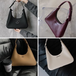 May include: Four leather shoulder bags in black, burgundy, tan, and cream. Each bag has a crescent shape with a single shoulder strap and a zipper closure. The bags are personalized with initials or names: "JB", "HERMIONA", "MIA", and "MONIKA".