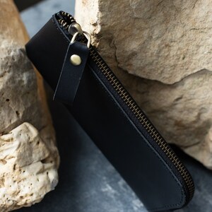May include: A black leather pen case with a brass zipper and a small leather pull tab. The case is closed and angled, resting against a light-colored stone surface.