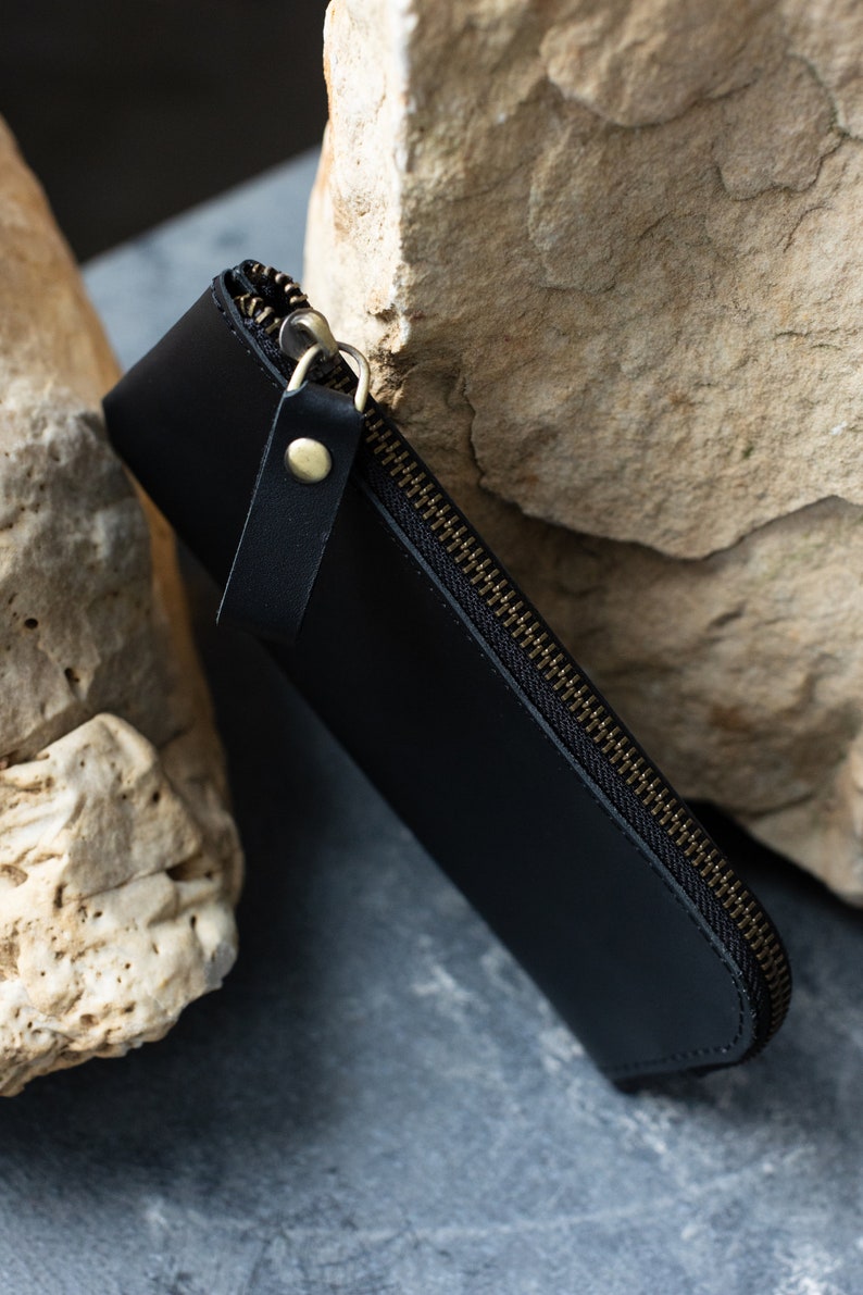 May include: A black leather pen case with a brass zipper and a small leather pull tab. The case is closed and angled, resting against a light-colored stone surface.