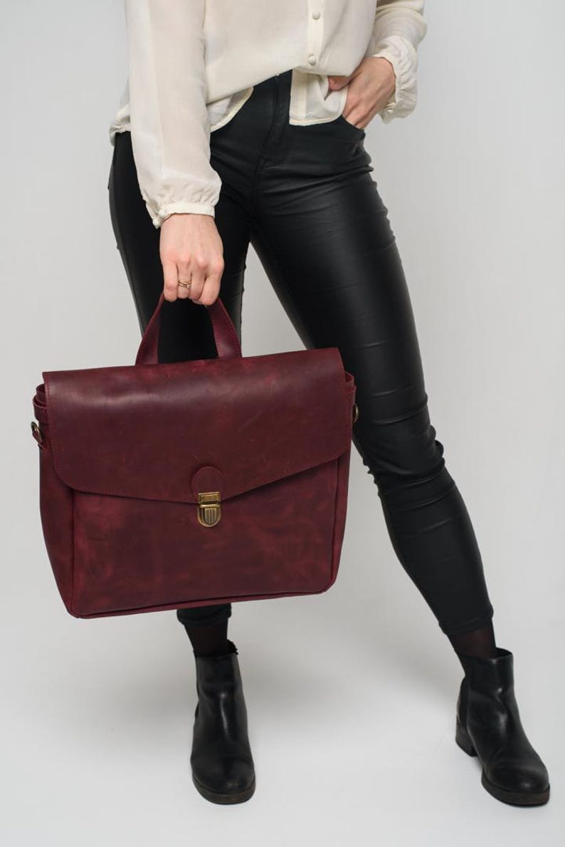 May include: A burgundy leather satchel with a top handle and a flap closure. The bag has a brass-coloured clasp and is held by a person wearing black trousers and boots. The background is plain white.