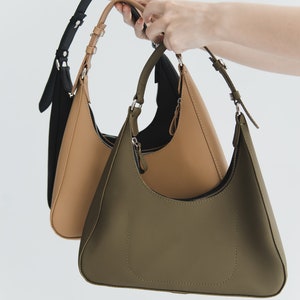 May include: Three shoulder bags in black, tan, and olive green are held up against a white background. Each bag has a curved, crescent shape with a zipper closure and adjustable strap. The bags are made of a smooth material.