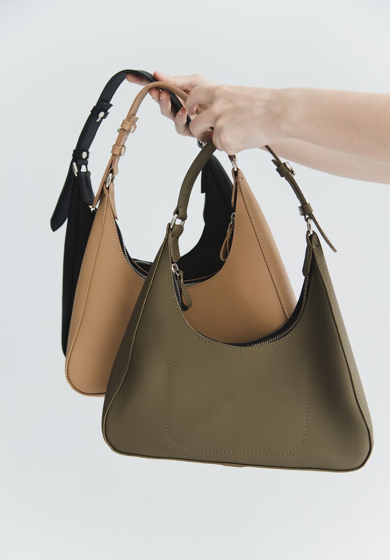 May include: Three shoulder bags in black, tan, and olive green are held up against a white background. Each bag has a curved, crescent shape with a zipper closure and adjustable strap. The bags are made of a smooth material.