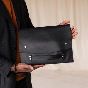 May include: A black leather portfolio with a flap closure and a handle. The portfolio has silver-coloured rivets and is held by a person wearing a dark grey blazer and a brown shirt.