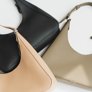 May include: Three leather shoulder bags in black, tan, and olive green are displayed against a white background. Each bag has a curved shape and a thin shoulder strap. The bags are made of smooth, shiny leather.