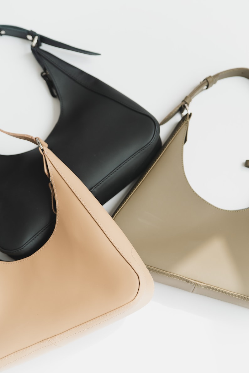 May include: Three leather shoulder bags in black, tan, and olive green are displayed against a white background. Each bag has a curved shape and a thin shoulder strap. The bags are made of smooth, shiny leather.