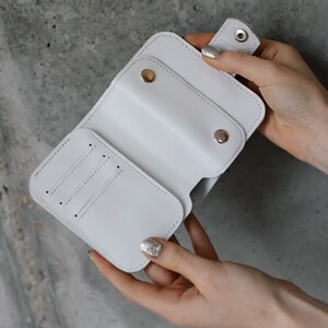 Pu&ograve; includere: Un portafoglio in pelle bianca con chiusura a scatto e pi&ugrave; fessure per carte. Il portafoglio &egrave; aperto, rivelando l'interno. Lo sfondo &egrave; grigio screziato.