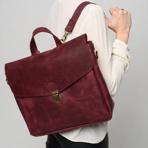 May include: A burgundy leather satchel with a flap closure and a gold-coloured clasp. The bag has a top handle and adjustable shoulder straps. The leather has a textured appearance. The bag is being carried over the shoulder.