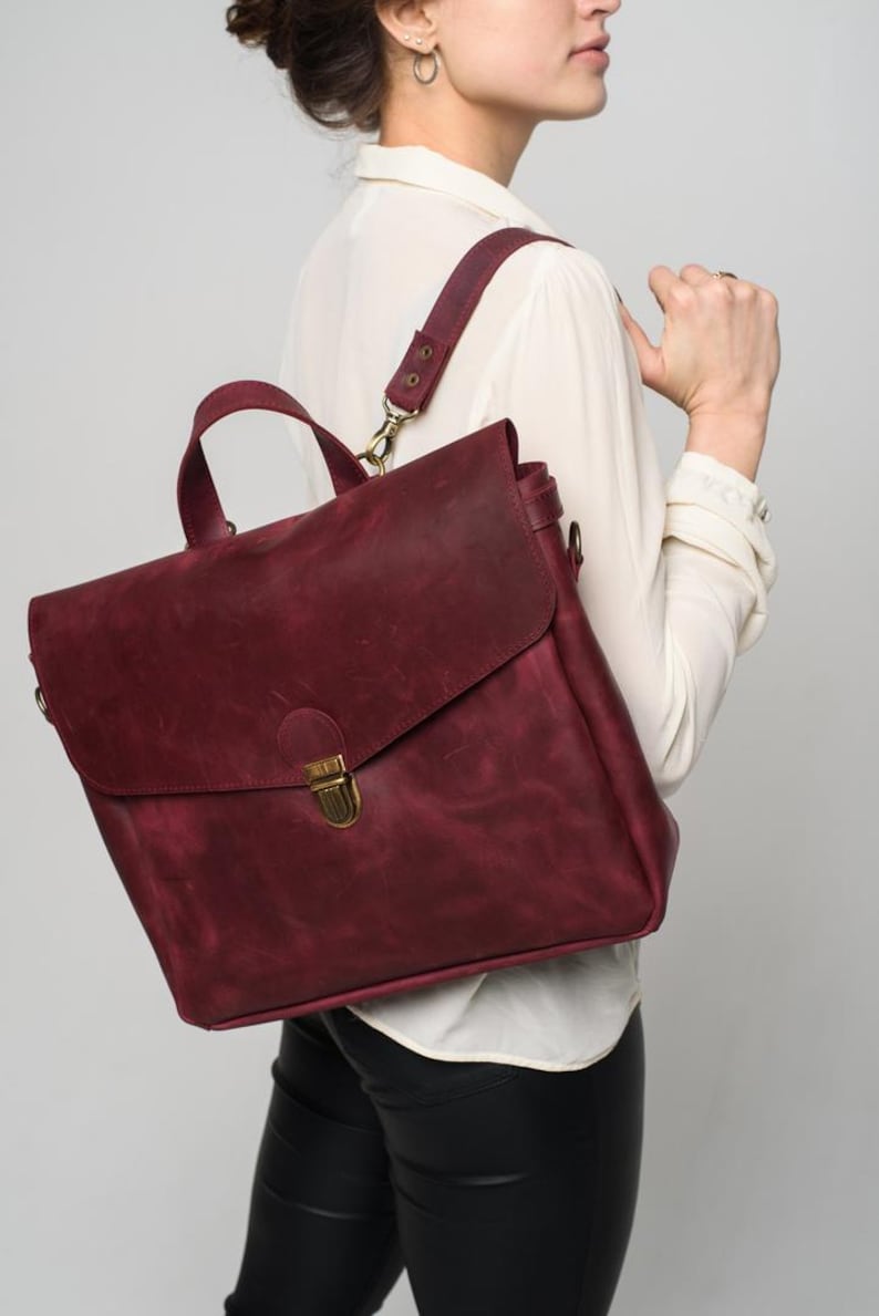 May include: A burgundy leather satchel with a flap closure and a gold-coloured clasp. The bag has a top handle and adjustable shoulder straps. The leather has a textured appearance. The bag is being carried over the shoulder.