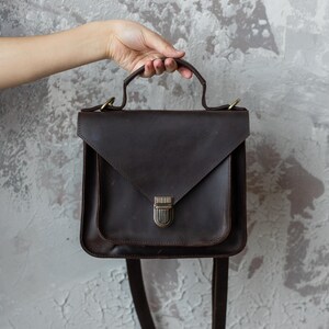 May include: A dark brown leather satchel with a flap closure and a top handle. The bag has a shoulder strap and a metal clasp. The bag is held by a hand against a textured gray background.