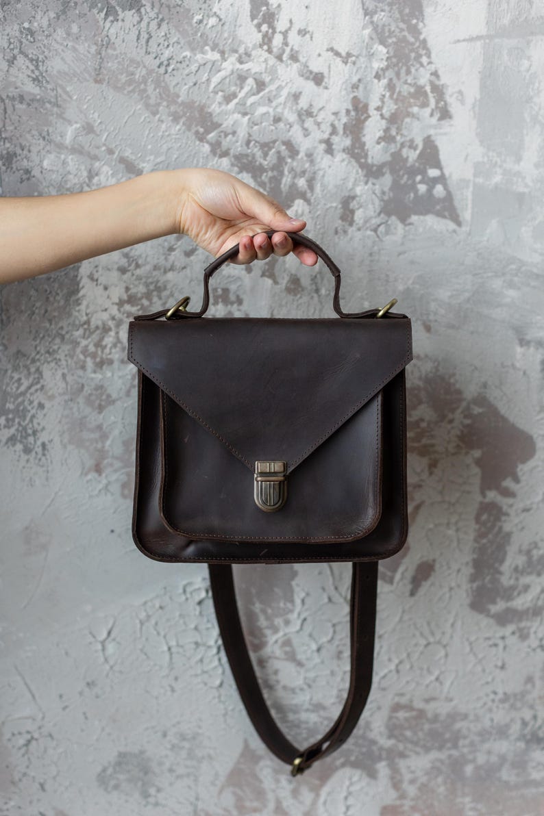 May include: A dark brown leather satchel with a flap closure and a top handle. The bag has a shoulder strap and a metal clasp. The bag is held by a hand against a textured gray background.