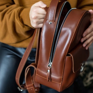 Peut inclure: Un sac &agrave; dos en cuir marron avec une fermeture &eacute;clair argent&eacute;e et une petite poche avant. Le sac a des bretelles r&eacute;glables et une poign&eacute;e sup&eacute;rieure. Le sac &agrave; dos est en cuir lisse et brillant.