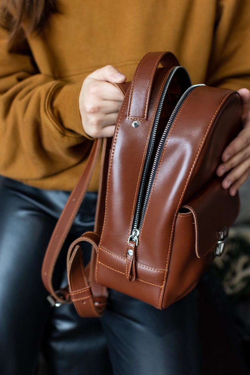 Peut inclure: Un sac &agrave; dos en cuir marron avec une fermeture &eacute;clair argent&eacute;e et une petite poche avant. Le sac a des bretelles r&eacute;glables et une poign&eacute;e sup&eacute;rieure. Le sac &agrave; dos est en cuir lisse et brillant.