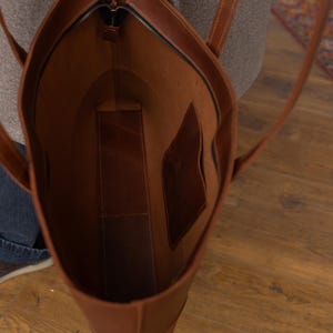 May include: A brown leather tote bag with a zipper closure. The bag has two long straps and an interior pocket. The bag is open, revealing the interior lining. The bag is made of a smooth, durable leather.