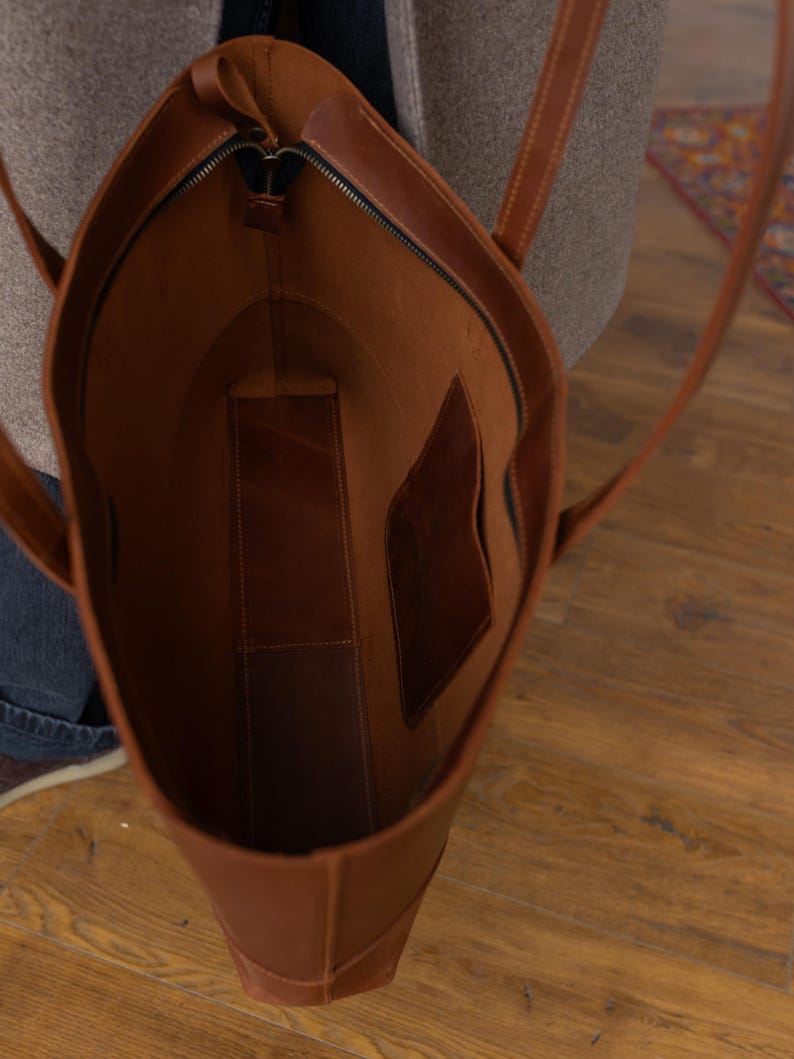 May include: A brown leather tote bag with a zipper closure. The bag has two long straps and an interior pocket. The bag is open, revealing the interior lining. The bag is made of a smooth, durable leather.