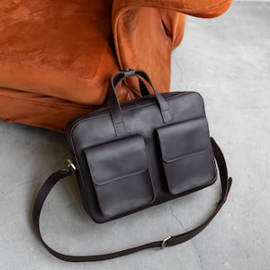 May include: A dark brown leather briefcase featuring two large front pockets with flap closures, a top zipper, two sturdy carrying handles, and an adjustable shoulder strap with metal hardware. The bag has a structured rectangular shape.