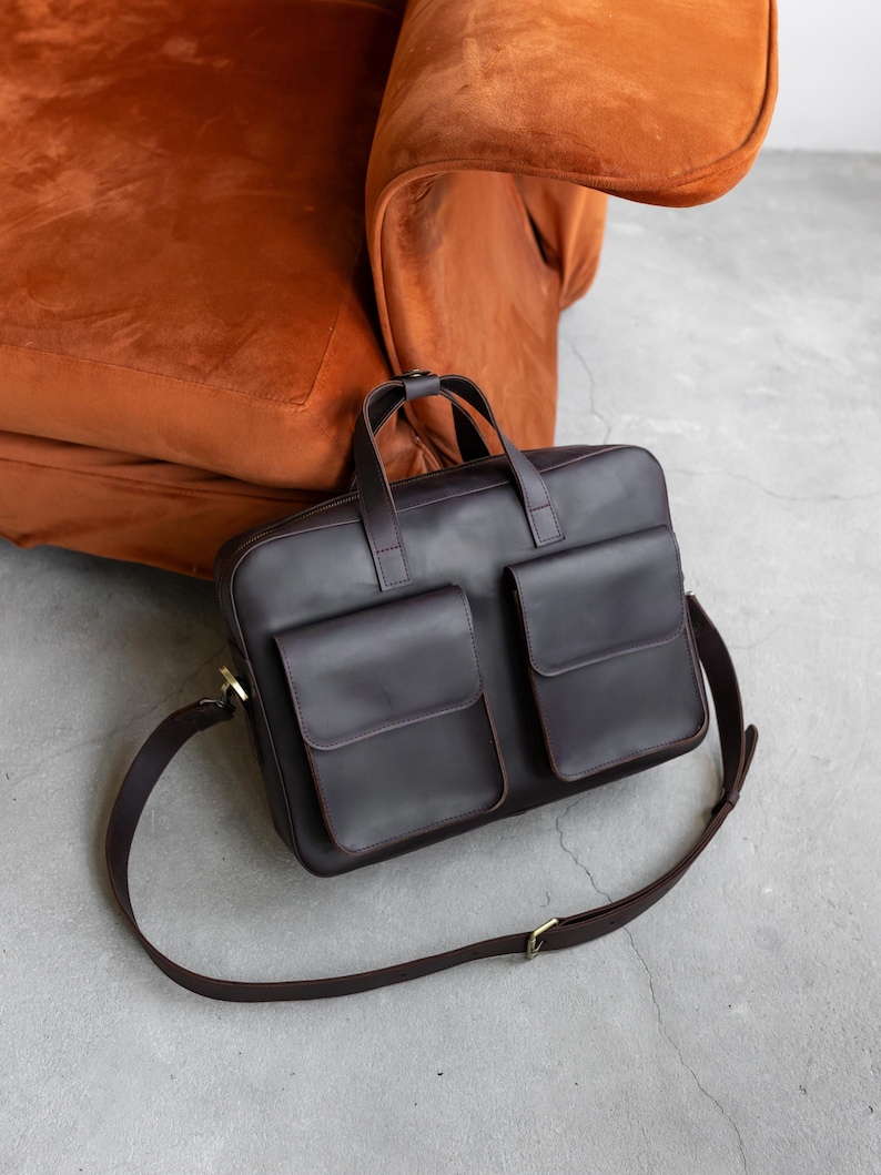 May include: A dark brown leather briefcase featuring two large front pockets with flap closures, a top zipper, two sturdy carrying handles, and an adjustable shoulder strap with metal hardware. The bag has a structured rectangular shape.