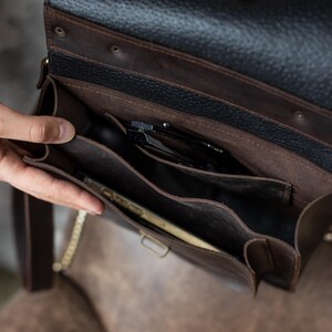 May include: A brown leather satchel with a black textured flap. The interior reveals multiple compartments, holding items like a phone and pens. The bag has a gold chain strap.