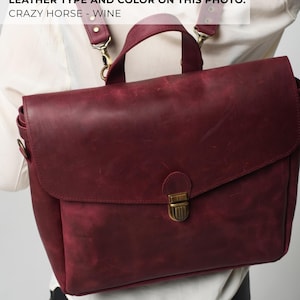 May include: A burgundy leather satchel with a flap closure and a top handle. The bag has adjustable shoulder straps and a brass-coloured clasp. The leather has a distressed appearance, and the image text reads "LEATHER TYPE AND COLOR ON THIS PHOTO: CRAZY HORSE - WINE."