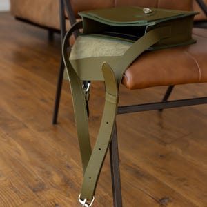 May include: Olive green leather crossbody bag with a flap closure and adjustable strap. The bag is open, revealing a glimpse of the interior. The strap hangs down, secured with a silver buckle. The bag rests on a brown leather chair.