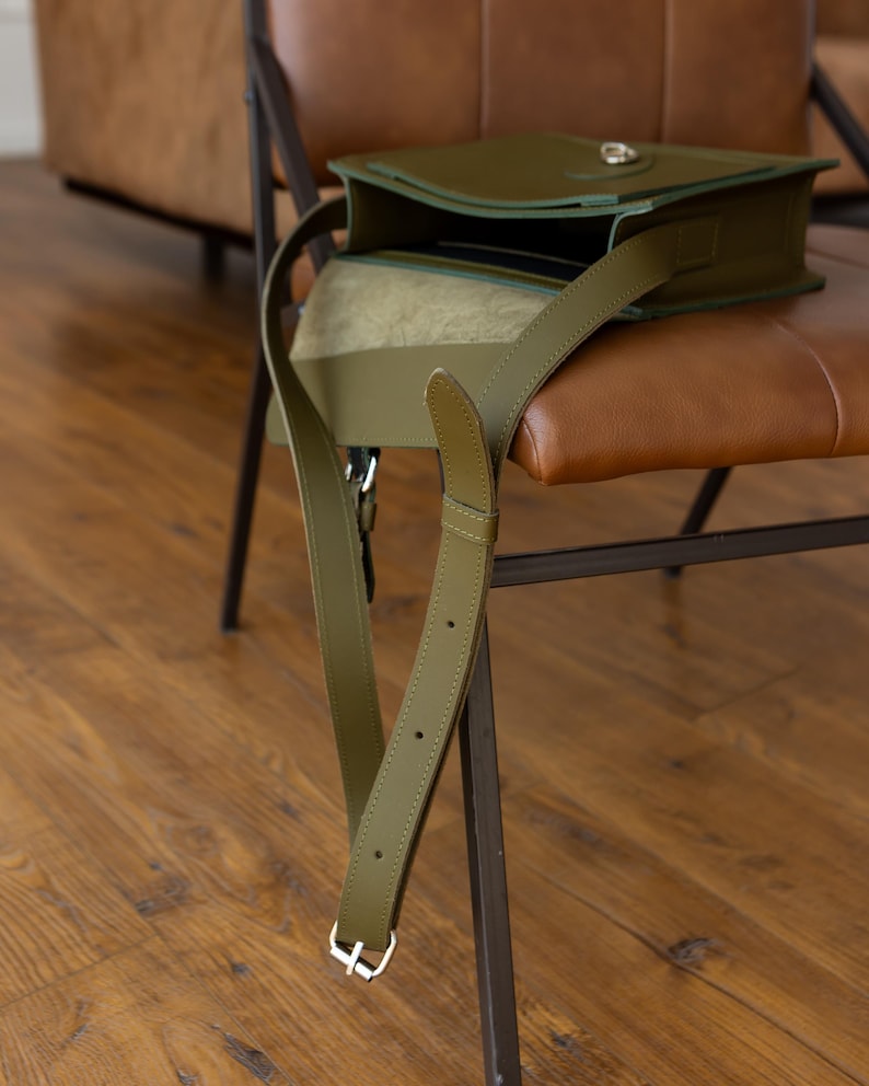 May include: Olive green leather crossbody bag with a flap closure and adjustable strap. The bag is open, revealing a glimpse of the interior. The strap hangs down, secured with a silver buckle. The bag rests on a brown leather chair.