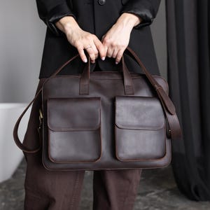 May include: A dark brown leather briefcase featuring a rectangular silhouette, two large front pockets with flap closures, two top handles, and an adjustable shoulder strap with metal hardware attachments on the sides of the bag.
